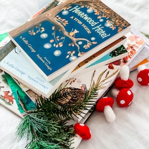Könnte beinhalten: Ein Stapel Kinderbücher mit farbenfrohen Einbänden, darunter "Heartwood Hotel: A True Home" von Kallie George. Dekorative rot-weiße Pilzornamente und ein Zweig grüner Kiefer sind ebenfalls vorhanden.
