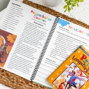 May include: An open cookbook with a spiral binding displays a recipe for "Pepparkakor Cookies." A colorful children's book titled "Pippi Longstocking" rests on the page. The image is set on a woven mat with a small potted plant.