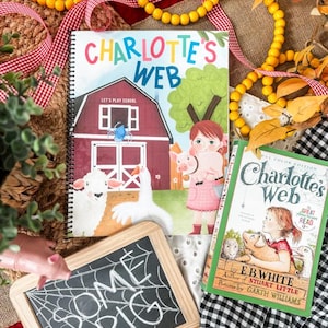 May include: Two copies of "Charlotte's Web" books, one spiral-bound and one paperback, with illustrations of farm animals. A small chalkboard with "SOME PIG" written on it is also present. The scene is decorated with yellow beads and red ribbon.
