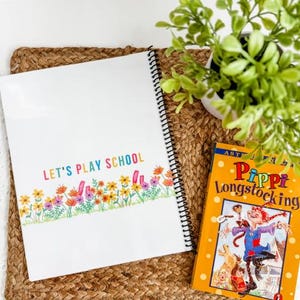May include: A spiral-bound notebook with the text "LET'S PLAY SCHOOL" in colorful letters, and a floral border. Next to it is a copy of "Pippi Longstocking". The items are on a woven mat with a small potted plant.