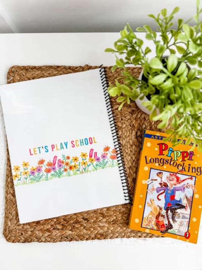 May include: A spiral-bound notebook with the text "LET'S PLAY SCHOOL" in colorful letters, and a floral border. Next to it is a copy of "Pippi Longstocking". The items are on a woven mat with a small potted plant.