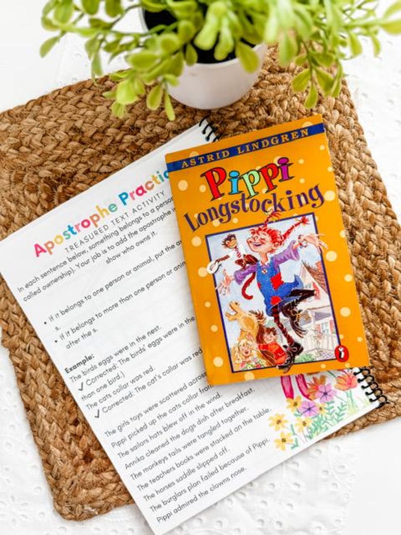 May include: A children's book titled "Pippi Longstocking" by Astrid Lindgren, featuring a vibrant illustration of Pippi on the cover. The book is placed on a woven mat alongside a worksheet about apostrophes. A small potted plant is in the background.
