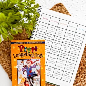 May include: A vibrant orange book titled "Pippi Longstocking" with a colorful illustration of the character. Next to it is a spiral-bound planner with a grid layout, featuring daily reading and activity prompts. The items rest on a woven mat.