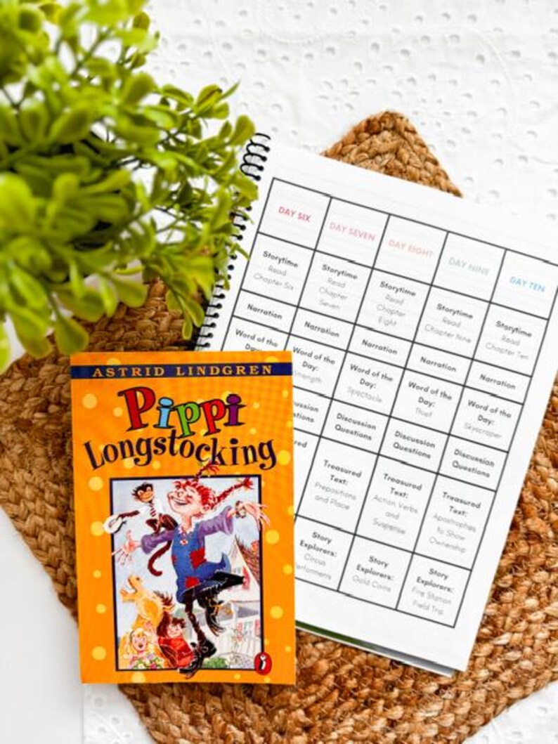 May include: A vibrant orange book titled "Pippi Longstocking" with a colorful illustration of the character. Next to it is a spiral-bound planner with a grid layout, featuring daily reading and activity prompts. The items rest on a woven mat.