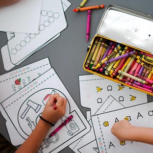 May include: Children's activity scene with coloring pages, crayons, and a yellow pencil box. The pages feature illustrations and the text "Let's Play Design". Crayons in various colors are scattered on the gray surface.