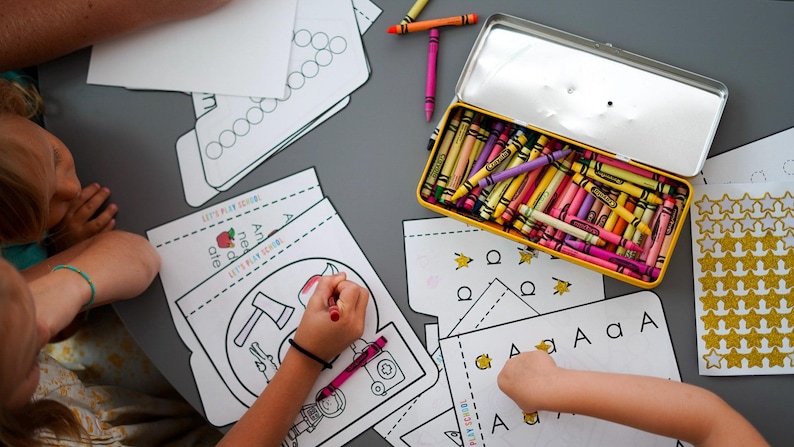 May include: Children's activity scene with coloring pages, crayons, and a yellow pencil box. The pages feature illustrations and the text "Let's Play Design". Crayons in various colors are scattered on the gray surface.