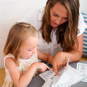 May include: A woman and a young child are seated at a grey table, engaged in an educational activity. They are looking at worksheets with the letter "A" and illustrations. A yellow crayon box and purple crayon are also on the table.