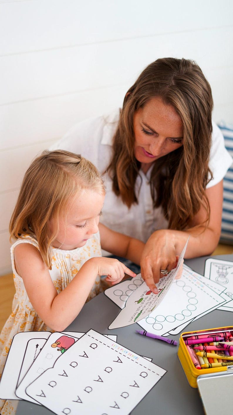 May include: A woman and a young child are seated at a gray table, engaged in an educational activity. They are looking at worksheets with the letter "A" and illustrations. A yellow crayon box and purple crayon are also on the table.