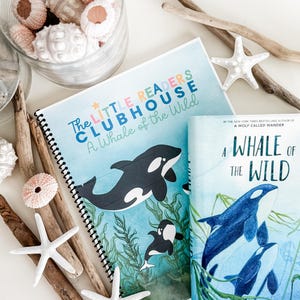 Puede incluir: Dos libros titulados "A Whale of the Wild" con ilustraciones de orcas. Uno está encuadernado en espiral y etiquetado como "The Little Readers Clubhouse". La imagen también presenta estrellas de mar, madera flotante y conchas de erizo de mar.