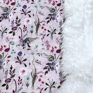 Puede incluir: Una manta suave de piel sint&eacute;tica blanca con un estampado floral en un lado. El estampado floral presenta una variedad de flores y hojas en tonos de rosa, morado y verde sobre un fondo morado claro.
