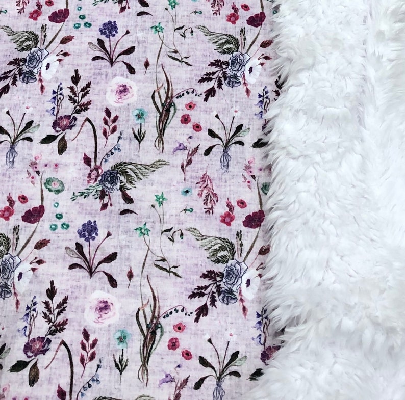 Puede incluir: Una manta suave de piel sint&eacute;tica blanca con un estampado floral en un lado. El estampado floral presenta una variedad de flores y hojas en tonos de rosa, morado y verde sobre un fondo morado claro.