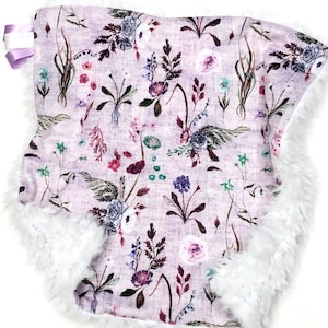 Puede incluir: Una manta de beb&eacute; suave y blanca de felpa con estampado floral lavanda. La manta tiene un ribete de piel sint&eacute;tica blanca alrededor de los bordes.