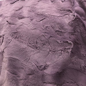 Puede incluir: Una manta suave y mullida de color morado claro. La manta tiene una textura peluda y un patr&oacute;n sutil.