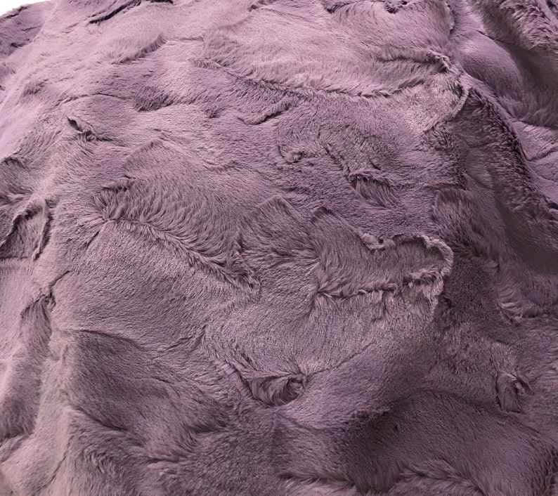 Puede incluir: Una manta suave y mullida de color morado claro. La manta tiene una textura peluda y un patr&oacute;n sutil.