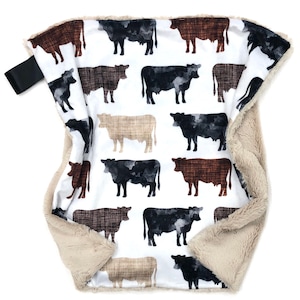 May include: A soft, rectangular security blanket featuring a white background with a repeating pattern of watercolor-style cows in shades of black, brown, and tan. The blanket has a plush, beige faux-fur backing and a small black fabric loop on one side.