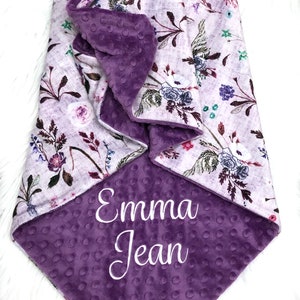 Puede incluir: Una manta para beb&eacute; de color morado y blanco con un estampado floral en un lado y una tela suave y texturizada de color morado en el otro lado. La manta est&aacute; personalizada con el nombre "Emma Jean" bordado en blanco.