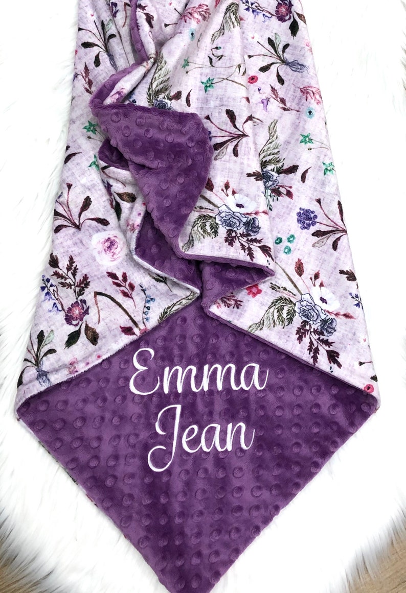 Puede incluir: Una manta para beb&eacute; de color morado y blanco con un estampado floral en un lado y una tela suave y texturizada de color morado en el otro lado. La manta est&aacute; personalizada con el nombre "Emma Jean" bordado en blanco.