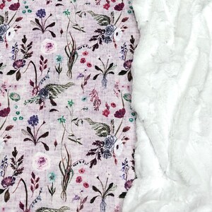 Puede incluir: Una manta de piel sint&eacute;tica blanca con un tejido estampado floral debajo. El tejido presenta una variedad de flores en tonos de rosa, morado y verde sobre un fondo morado claro.