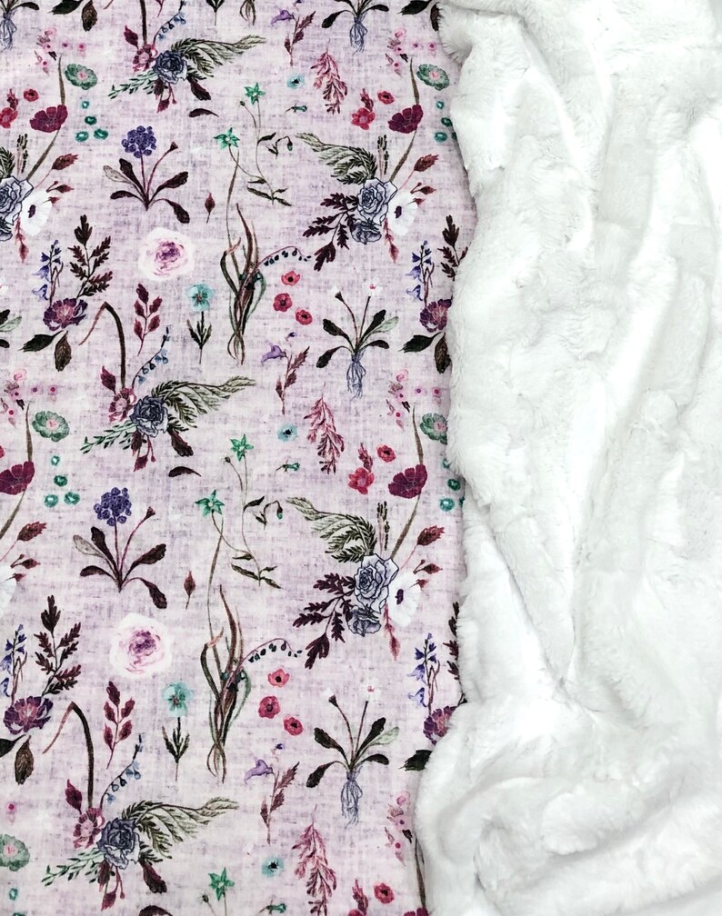 Puede incluir: Una manta de piel sint&eacute;tica blanca con un tejido estampado floral debajo. El tejido presenta una variedad de flores en tonos de rosa, morado y verde sobre un fondo morado claro.