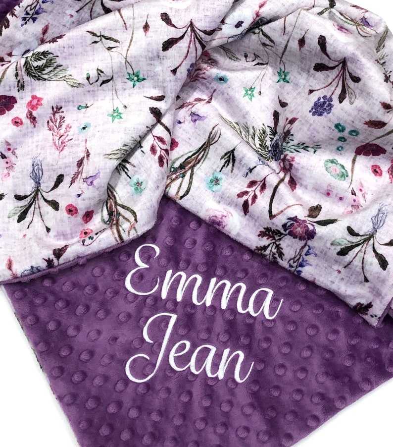 Puede incluir: Manta de beb&eacute; de color morado y blanco con un patr&oacute;n floral en un lado y una tela minky morada en el otro lado. El lado minky tiene el nombre "Emma Jean" bordado en blanco.