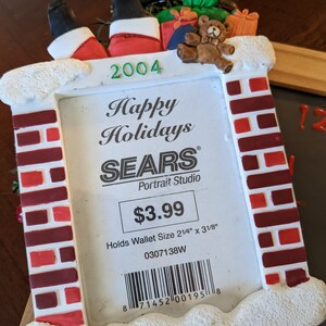 May include: A Christmas-themed photo frame with a chimney design. The frame features Santa's legs, gifts, and a teddy bear. The text "Happy Holidays" and "SEARS Portrait Studio" are visible. The frame holds a wallet-sized photo, 2 1/4" x 3 1/8".