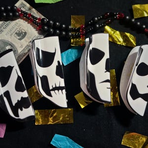 May include: Four white and black skull-shaped keyrings with a black and white skull design. The keyrings are on a black surface with gold and blue accents.
