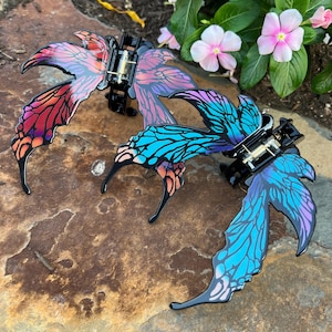 May include: Two butterfly wing hair clips with black plastic hinges and metal springs. One clip features a gradient of red, orange, and purple wing patterns. The second clip displays a gradient of blue, teal, and purple wing patterns with black veins.
