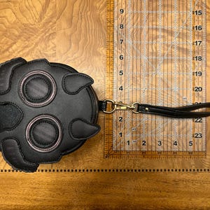 K&ouml;nnte beinhalten: Schwarze, runde Leder-Clutch mit einem einzigartigen Design, das zwei gro&szlig;e augen&auml;hnliche Kreise und zwei kleinere Kreise aufweist, die alle rosa umrandet sind. Sie hat eine Handschlaufe und einen goldfarbenen Verschluss. Die Clutch hat einen Durchmesser von etwa 15 cm.