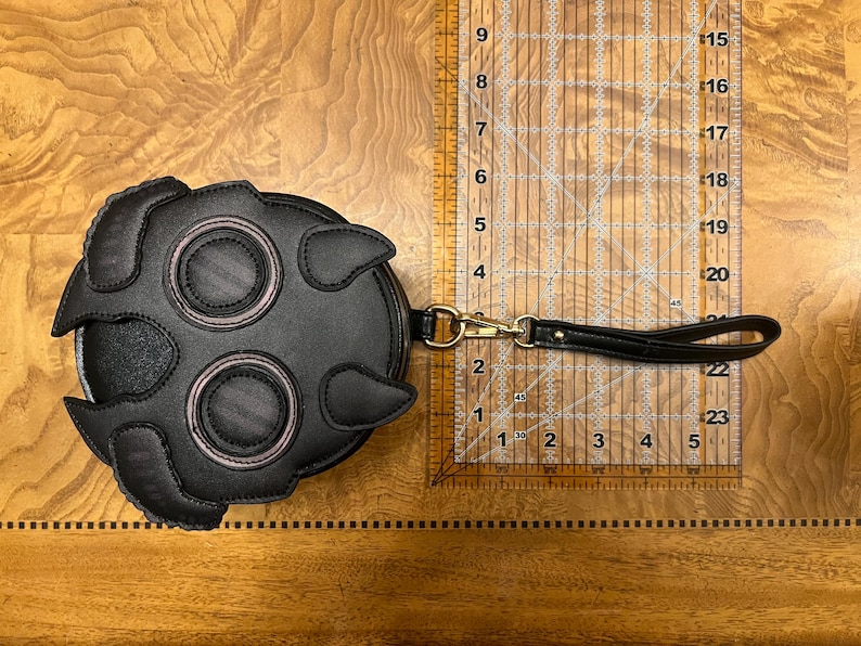 K&ouml;nnte beinhalten: Schwarze, runde Leder-Clutch mit einem einzigartigen Design, das zwei gro&szlig;e augen&auml;hnliche Kreise und zwei kleinere Kreise aufweist, die alle rosa umrandet sind. Sie hat eine Handschlaufe und einen goldfarbenen Verschluss. Die Clutch hat einen Durchmesser von etwa 15 cm.