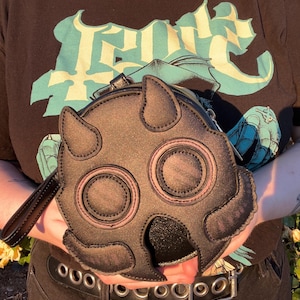 K&ouml;nnte beinhalten: Schwarze, runde Handtasche mit einem einzigartigen, stilisierten Design, das einer Maske oder einem Gesicht &auml;hnelt. Die Tasche hat zwei gro&szlig;e kreisf&ouml;rmige Augen, spitze, ohrartige Vorspr&uuml;nge und einen kleinen Griff. Die Tasche wird vor einem schwarzen T-Shirt-Hintergrund gehalten.