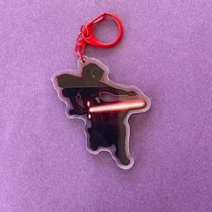 May include: A clear acrylic keychain featuring a black silhouette of a character wielding a red lightsaber. The keychain has a red metal clasp. The design is outlined in red and is on a purple background.
