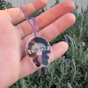 May include: A clear acrylic charm with a cartoon illustration of a character with black hair and a purple cape. The charm is attached to a purple ball chain. The background is blurred.