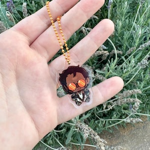 May include: A clear acrylic charm featuring a cartoon character with dark hair and orange-tinted circular glasses, wearing a white and black outfit. The charm is attached to a metallic orange ball chain, creating a small decorative accessory.