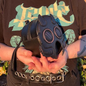 K&ouml;nnte beinhalten: Schwarze, runde Rei&szlig;verschlusstasche mit Gasmasken-Design. Die Tasche hat einen Griff und verf&uuml;gt &uuml;ber ein Schwarz- und Pink-Farbschema. Das Gasmasken-Design umfasst zwei kreisf&ouml;rmige Augen&ouml;ffnungen und einen detaillierten Nasen- und Mundbereich.