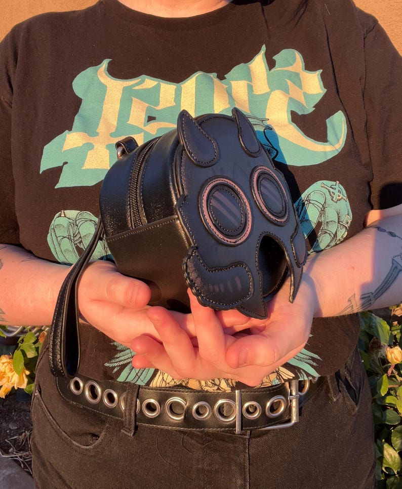 K&ouml;nnte beinhalten: Schwarze, runde Rei&szlig;verschlusstasche mit Gasmasken-Design. Die Tasche hat einen Griff und verf&uuml;gt &uuml;ber ein Schwarz- und Pink-Farbschema. Das Gasmasken-Design umfasst zwei kreisf&ouml;rmige Augen&ouml;ffnungen und einen detaillierten Nasen- und Mundbereich.