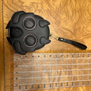 K&ouml;nnte beinhalten: Schwarze runde Handtasche mit einem einzigartigen Design mit zwei gro&szlig;en augenartigen Kreisen mit rosa Akzenten und einem Griff. Die Handtasche hat einen Durchmesser von etwa 15 cm.