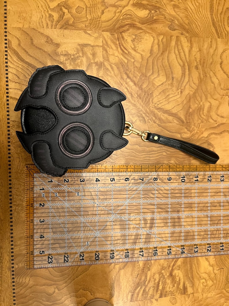 K&ouml;nnte beinhalten: Schwarze runde Handtasche mit einem einzigartigen Design mit zwei gro&szlig;en augenartigen Kreisen mit rosa Akzenten und einem Griff. Die Handtasche hat einen Durchmesser von etwa 15 cm.