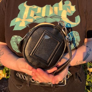 K&ouml;nnte beinhalten: Schwarze, runde Handtasche mit einem transparenten, rechteckigen Fenster auf der Vorderseite. Die Tasche hat einen kleinen Griff und eine silberne Kette. Die Person tr&auml;gt ein schwarzes T-Shirt mit einem t&uuml;rkis-gelben Grafikdesign.