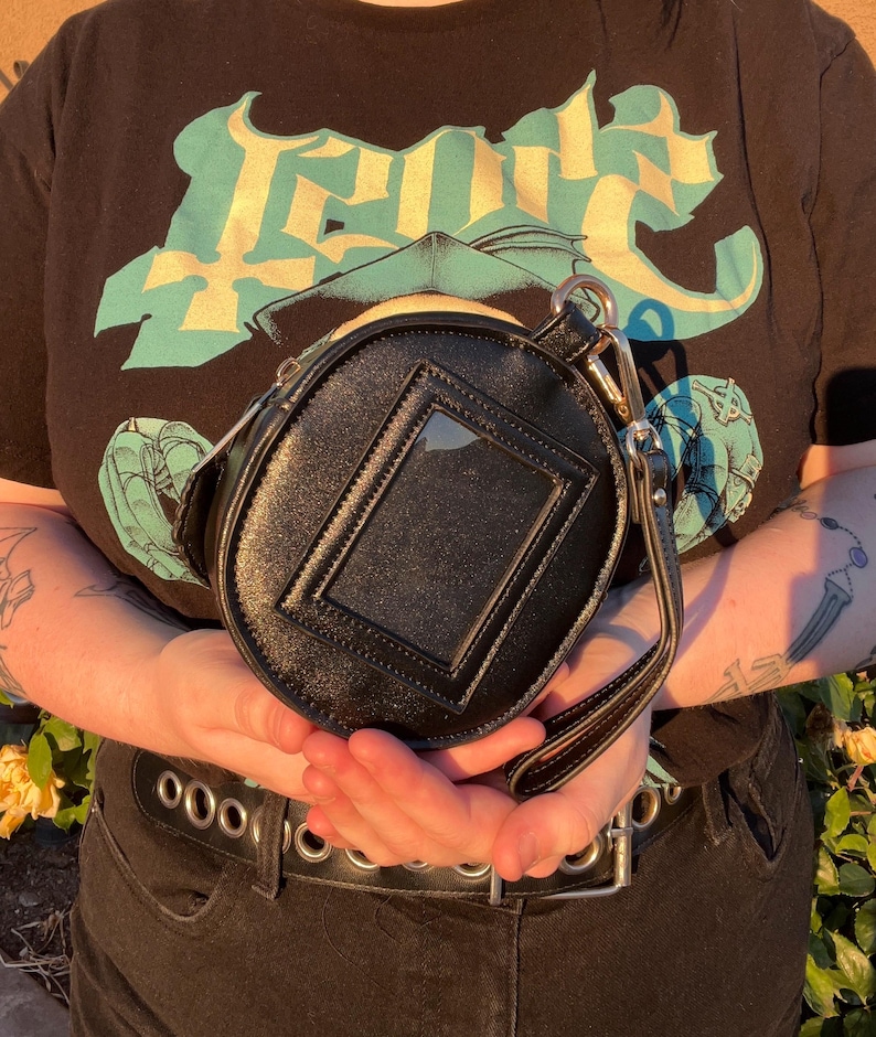 K&ouml;nnte beinhalten: Schwarze, runde Handtasche mit einem transparenten, rechteckigen Fenster auf der Vorderseite. Die Tasche hat einen kleinen Griff und eine silberne Kette. Die Person tr&auml;gt ein schwarzes T-Shirt mit einem t&uuml;rkis-gelben Grafikdesign.
