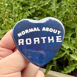 May include: A heart-shaped pin with a glossy dark blue marbled background. The text "NORMAL ABOUT ROATHE" is printed in white, with the word "ROATHE" featuring a stylized, glitch-like font effect across the center of the blue surface.
