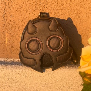 K&ouml;nnte beinhalten: Eine dunkelbraune, runde Handtasche mit einem einzigartigen Masken-Design. Die Tasche hat zwei gro&szlig;e, augen&auml;hnliche Kreise mit rosa Akzenten, zwei hornartige Formen oben und einen kleinen Griff. Die Tasche steht vor einer strukturierten, orangefarbenen Wand.