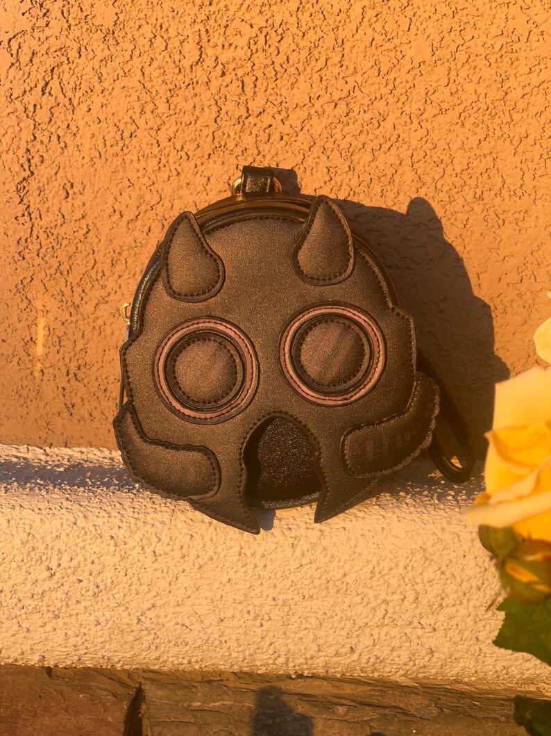 K&ouml;nnte beinhalten: Eine dunkelbraune, runde Handtasche mit einem einzigartigen Masken-Design. Die Tasche hat zwei gro&szlig;e, augen&auml;hnliche Kreise mit rosa Akzenten, zwei hornartige Formen oben und einen kleinen Griff. Die Tasche steht vor einer strukturierten, orangefarbenen Wand.