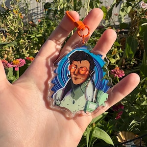 Puede incluir: Un llavero acrílico transparente con una ilustración estilizada de una persona con gafas de color naranja y auriculares. El llavero tiene un cierre naranja y un fondo azul y blanco. La persona lleva un atuendo verde claro.