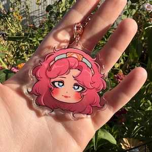 Puede incluir: Un llavero acrílico transparente con una ilustración de dibujos animados de un personaje con cabello rizado rosa, ojos azules y una diadema. El llavero tiene un cierre de oro rosa y se sostiene en una mano sobre un fondo de flores.