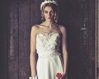 Op de afbeelding: Een bruid in een witte strapless bruidsjurk met een bloemencorsage en een enkele rode roos. De jurk heeft een zwierige rok en de bruid staat voor een houten deur.