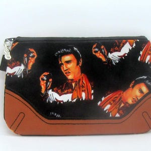 May include: Black zippered pouch featuring multiple images of Elvis Presley in performance attire. The pouch has a brown bottom and a small guitar-shaped zipper pull. The background is black.