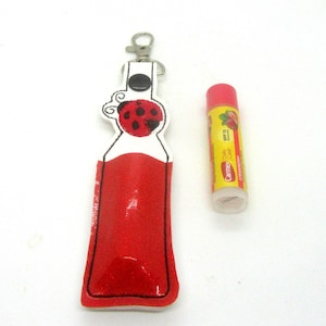 Könnte beinhalten: Ein roter Marienkäfer-Lippenbalsamhalter mit weißem Hintergrund und einem silbernen Schlüsselanhänger. Der Halter ist so konzipiert, dass er in ein Lippenbalsamröhrchen in Standardgröße passt. Der Lippenbalsamhalter wird neben einer Tube Carmex Lippenbalsam mit SPF 15 und Erdbeergeschmack gezeigt.
