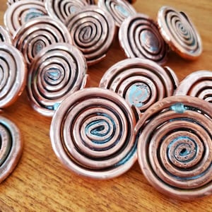 Peut inclure: Collection d'objets décoratifs en forme de spirale de couleur cuivrée. Chaque pièce présente un motif en spirale concentrique en relief avec des reflets de patine bleu-vert. Les objets sont disposés sur une surface en bois.