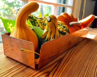 May include: A copper tray filled with gourds and squash in various shades of orange, yellow, and green. The tray is sitting on a wooden surface.