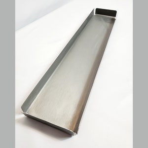 May include: A long, rectangular metal tray with a brushed finish. The tray has a slight curve at the top and bottom edges.